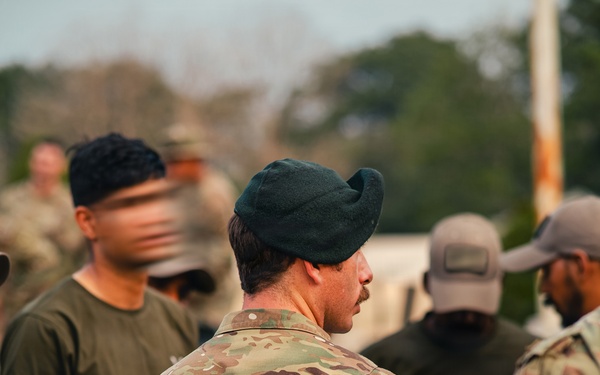 A group of military personnel gathered outdoors, with the central figure wearing OCP (Operational Camouflage Pattern) uniform and a dark green beret. Multiple individuals in various military and tactical attire are visible, suggesting a formation, briefing, or assembly event. The setting appears to be a military installation or training area with trees visible in the background.