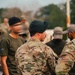 A group of military personnel gathered outdoors, with the central figure wearing OCP (Operational Camouflage Pattern) uniform and a dark green beret. Multiple individuals in various military and tactical attire are visible, suggesting a formation, briefing, or assembly event. The setting appears to be a military installation or training area with trees visible in the background.
