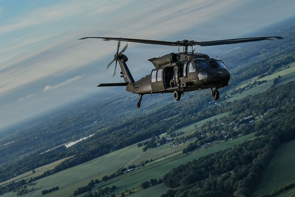 A military helicopter, identifiable as a UH-60 Black Hawk, is captured in flight at altitude over a rural landscape featuring green fields, forests, and scattered buildings. The aircraft is painted in a dark olive/black military finish and is photographed from another airborne platform. The image shows a wide aerial perspective with a partly cloudy sky in the background.