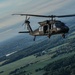 A military helicopter, identifiable as a UH-60 Black Hawk, is captured in flight at altitude over a rural landscape featuring green fields, forests, and scattered buildings. The aircraft is painted in a dark olive/black military finish and is photographed from another airborne platform. The image shows a wide aerial perspective with a partly cloudy sky in the background.