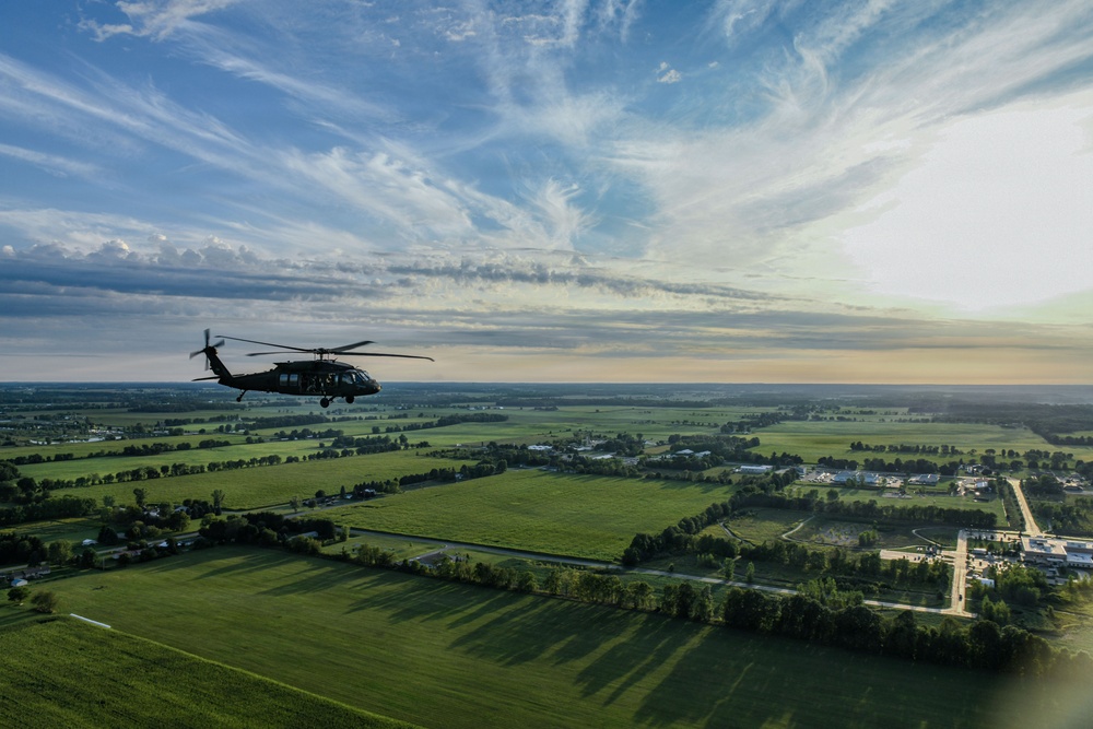 An aerial photograph captures a military helicopter, consistent with a UH-60 Black Hawk, flying over a flat, rural landscape of green fields and scattered tree lines. The image appears to be taken from another aircraft flying in formation or proximity. The scene is bathed in warm late-afternoon or early-evening light beneath a partly cloudy sky with wispy cirrus clouds.