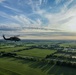 An aerial photograph captures a military helicopter, consistent with a UH-60 Black Hawk, flying over a flat, rural landscape of green fields and scattered tree lines. The image appears to be taken from another aircraft flying in formation or proximity. The scene is bathed in warm late-afternoon or early-evening light beneath a partly cloudy sky with wispy cirrus clouds.