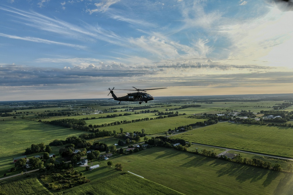 A military helicopter, consistent with a UH-60 Black Hawk, is captured in flight over a flat, rural agricultural landscape. The aerial perspective reveals a patchwork of green fields, scattered farmsteads, tree lines, and rural roads stretching to the horizon. The image was taken from another airborne platform during what appears to be a late afternoon or early evening flight.