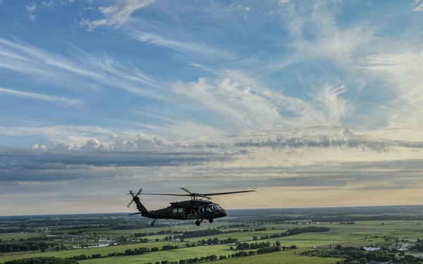 A military helicopter, consistent with a UH-60 Black Hawk, is captured in flight over a flat, rural agricultural landscape. The aerial perspective reveals a patchwork of green fields, scattered farmsteads, tree lines, and rural roads stretching to the horizon. The image was taken from another airborne platform during what appears to be a late afternoon or early evening flight.
