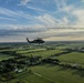 A military helicopter, consistent with a UH-60 Black Hawk, is captured in flight over a flat, rural agricultural landscape. The aerial perspective reveals a patchwork of green fields, scattered farmsteads, tree lines, and rural roads stretching to the horizon. The image was taken from another airborne platform during what appears to be a late afternoon or early evening flight.