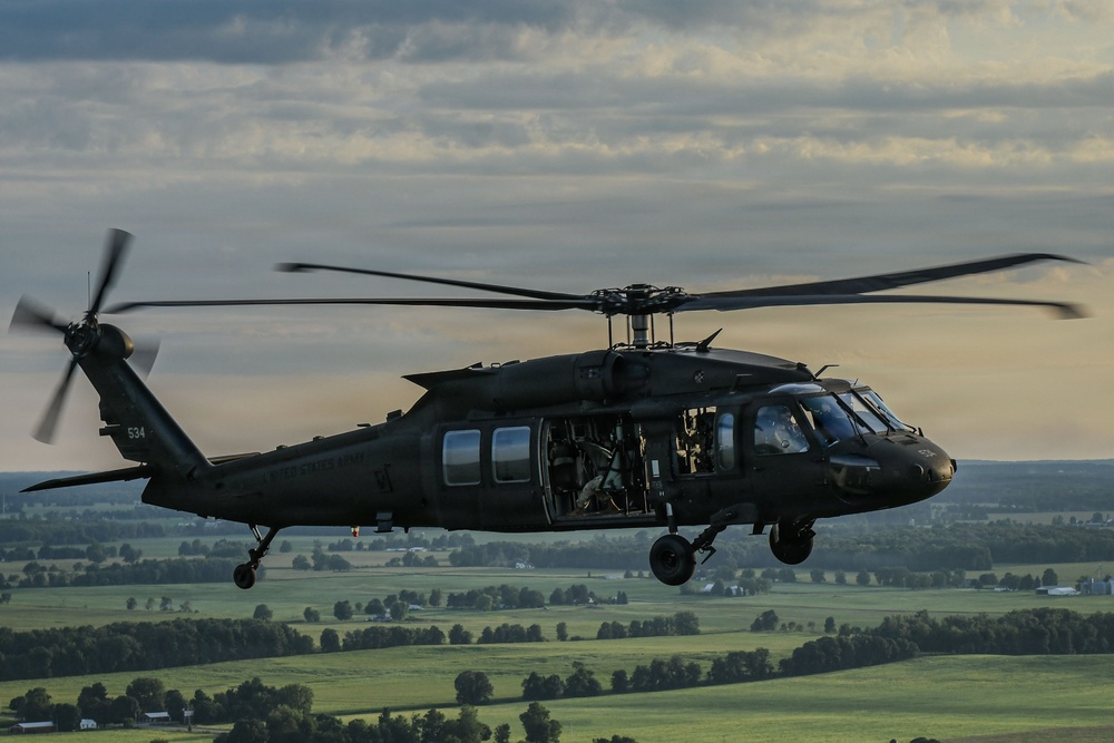 A military utility helicopter, consistent with a UH-60 Black Hawk, is captured in flight over a rural, green landscape at low altitude during overcast conditions. The aircraft is painted in flat olive/dark green military livery and has its side door open with personnel visible inside. The image is a high-quality aerial photograph likely taken from another aircraft flying in formation.