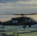 A military utility helicopter, consistent with a UH-60 Black Hawk, is captured in flight over a rural, green landscape at low altitude during overcast conditions. The aircraft is painted in flat olive/dark green military livery and has its side door open with personnel visible inside. The image is a high-quality aerial photograph likely taken from another aircraft flying in formation.