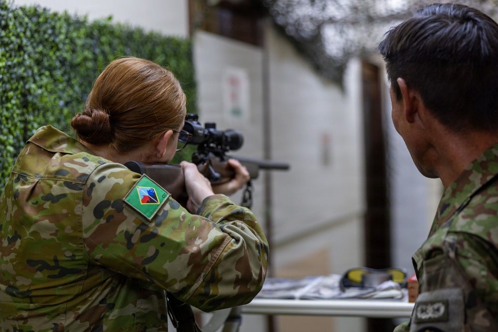 A female soldier in multicam camouflage uniform is aiming a scoped rifle at a shooting range or training facility. A male soldier in similar uniform observes from the right side. The patch on the female soldier's left shoulder appears to be a Samoa or Pacific Island nation flag patch, suggesting a multinational military training exercise.