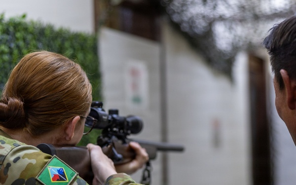 A female soldier in multicam camouflage uniform is aiming a scoped rifle at a shooting range or training facility. A male soldier in similar uniform observes from the right side. The patch on the female soldier's left shoulder appears to be a Samoa or Pacific Island nation flag patch, suggesting a multinational military training exercise.