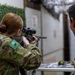 A female soldier in multicam camouflage uniform is aiming a scoped rifle at a shooting range or training facility. A male soldier in similar uniform observes from the right side. The patch on the female soldier's left shoulder appears to be a Samoa or Pacific Island nation flag patch, suggesting a multinational military training exercise.