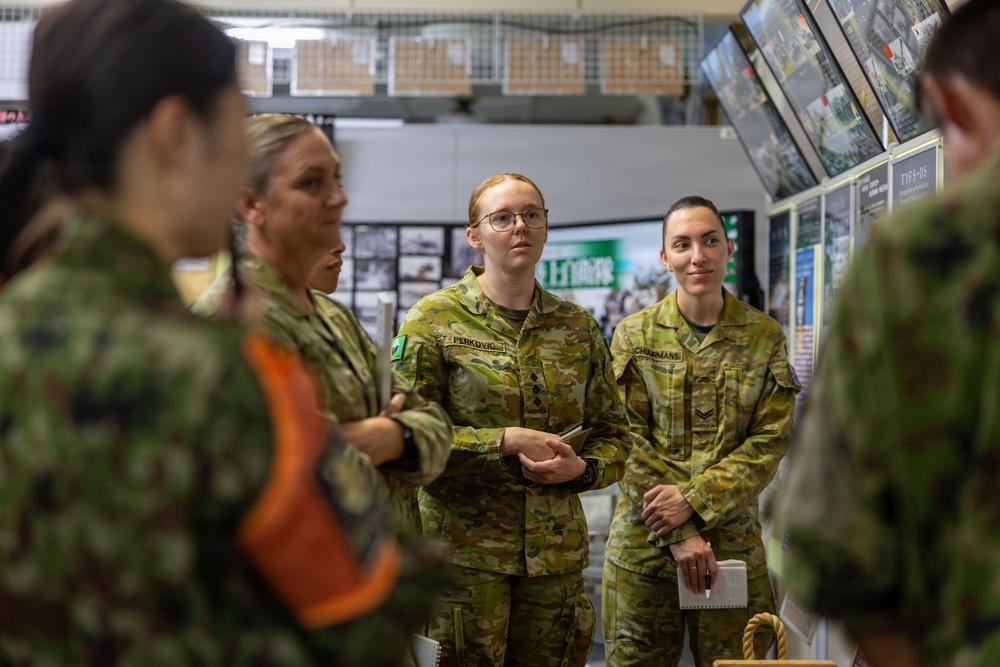A group of Australian Defence Force personnel, predominantly female, are gathered indoors in what appears to be a briefing or tour environment. They are wearing Australian Multicam Camouflage (AMCU) uniforms with visible name tags and rank insignia. The setting includes display screens, informational boards, and what appears to be a military facility or exhibition space with Japanese text visible in the background.