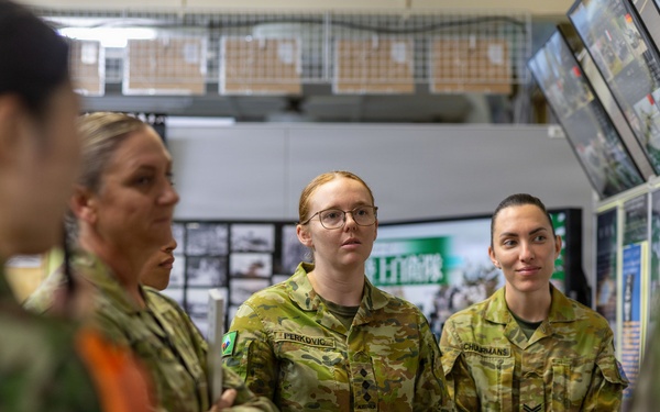 A group of Australian Defence Force personnel, predominantly female, are gathered indoors in what appears to be a briefing or tour environment. They are wearing Australian Multicam Camouflage (AMCU) uniforms with visible name tags and rank insignia. The setting includes display screens, informational boards, and what appears to be a military facility or exhibition space with Japanese text visible in the background.