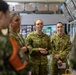 A group of Australian Defence Force personnel, predominantly female, are gathered indoors in what appears to be a briefing or tour environment. They are wearing Australian Multicam Camouflage (AMCU) uniforms with visible name tags and rank insignia. The setting includes display screens, informational boards, and what appears to be a military facility or exhibition space with Japanese text visible in the background.