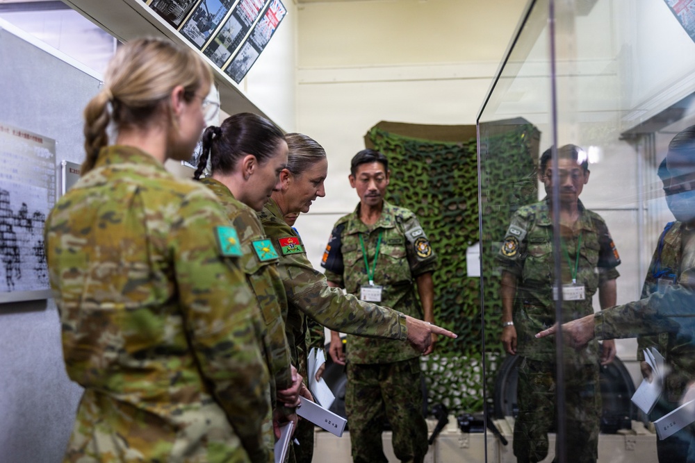 A group of military personnel from at least two different nations are gathered in what appears to be a briefing room or display area. Australian Army soldiers (identifiable by their AUSCAM uniforms and shoulder patches) are interacting with Japanese Ground Self-Defense Force members wearing green camouflage uniforms. One Australian officer is pointing at a glass display case, suggesting a tour or information exchange is taking place.