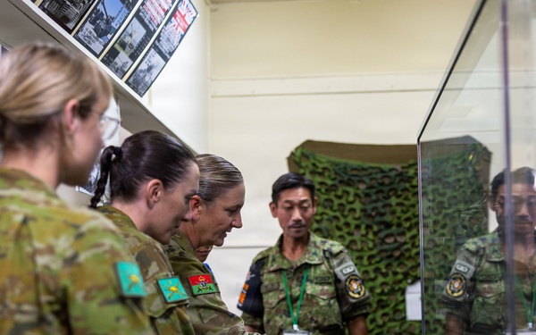 A group of military personnel from at least two different nations are gathered in what appears to be a briefing room or display area. Australian Army soldiers (identifiable by their AUSCAM uniforms and shoulder patches) are interacting with Japanese Ground Self-Defense Force members wearing green camouflage uniforms. One Australian officer is pointing at a glass display case, suggesting a tour or information exchange is taking place.