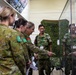 A group of military personnel from at least two different nations are gathered in what appears to be a briefing room or display area. Australian Army soldiers (identifiable by their AUSCAM uniforms and shoulder patches) are interacting with Japanese Ground Self-Defense Force members wearing green camouflage uniforms. One Australian officer is pointing at a glass display case, suggesting a tour or information exchange is taking place.