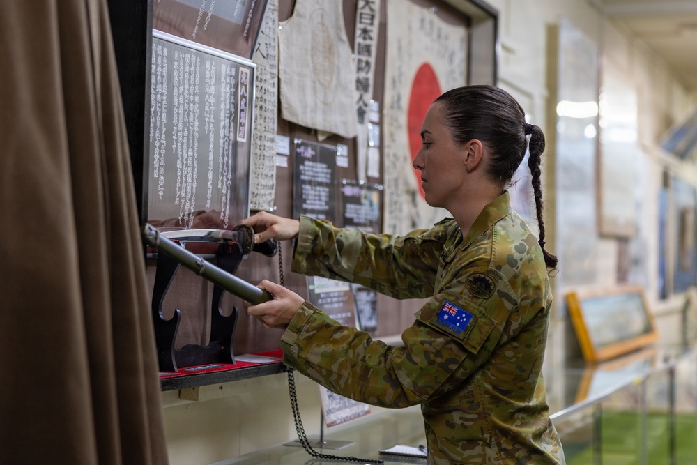A female Australian Defence Force (ADF) soldier in MultiCam camouflage uniform is examining a Japanese sword (katana) on display at what appears to be a military or historical museum. The exhibit features Japanese wartime artifacts including documents written in Japanese kanji, a Japanese national flag (Hinomaru), and what appears to be a traditional garment. The setting suggests a cultural or historical exchange visit to a Japanese military or heritage facility.