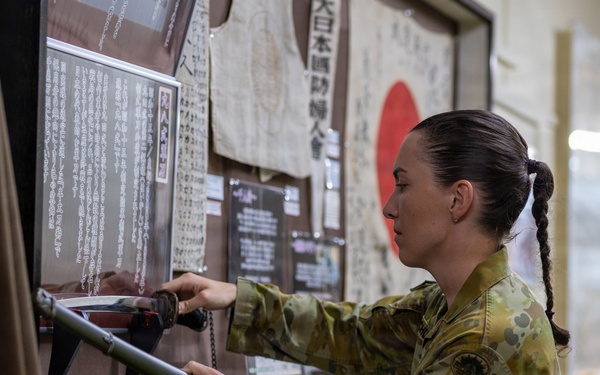 A female Australian Defence Force (ADF) soldier in MultiCam camouflage uniform is examining a Japanese sword (katana) on display at what appears to be a military or historical museum. The exhibit features Japanese wartime artifacts including documents written in Japanese kanji, a Japanese national flag (Hinomaru), and what appears to be a traditional garment. The setting suggests a cultural or historical exchange visit to a Japanese military or heritage facility.
