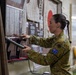 A female Australian Defence Force (ADF) soldier in MultiCam camouflage uniform is examining a Japanese sword (katana) on display at what appears to be a military or historical museum. The exhibit features Japanese wartime artifacts including documents written in Japanese kanji, a Japanese national flag (Hinomaru), and what appears to be a traditional garment. The setting suggests a cultural or historical exchange visit to a Japanese military or heritage facility.