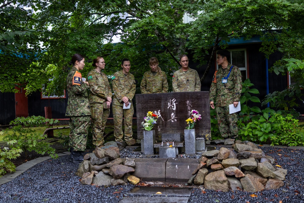 A group of six military personnel, appearing to include both Japanese Ground Self-Defense Force members and Australian Army soldiers, are gathered around a memorial stone monument with Japanese kanji inscriptions and flower offerings. The scene appears to be a solemn commemorative visit at what looks like a military installation or memorial garden in Japan. The monument is surrounded by rocks and small stone pedestals holding flowers and candles.