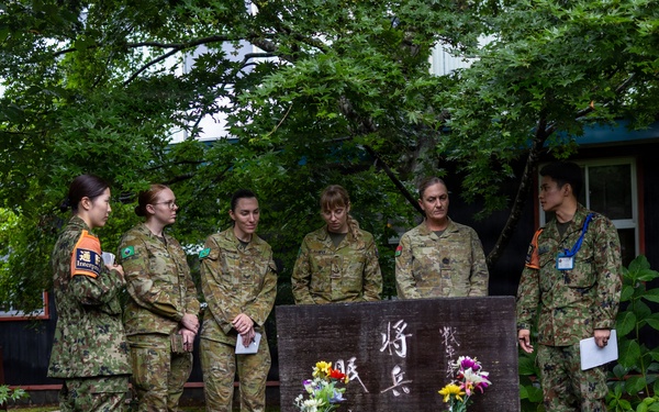 A group of six military personnel, appearing to include both Japanese Ground Self-Defense Force members and Australian Army soldiers, are gathered around a memorial stone monument with Japanese kanji inscriptions and flower offerings. The scene appears to be a solemn commemorative visit at what looks like a military installation or memorial garden in Japan. The monument is surrounded by rocks and small stone pedestals holding flowers and candles.