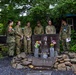 A group of six military personnel, appearing to include both Japanese Ground Self-Defense Force members and Australian Army soldiers, are gathered around a memorial stone monument with Japanese kanji inscriptions and flower offerings. The scene appears to be a solemn commemorative visit at what looks like a military installation or memorial garden in Japan. The monument is surrounded by rocks and small stone pedestals holding flowers and candles.