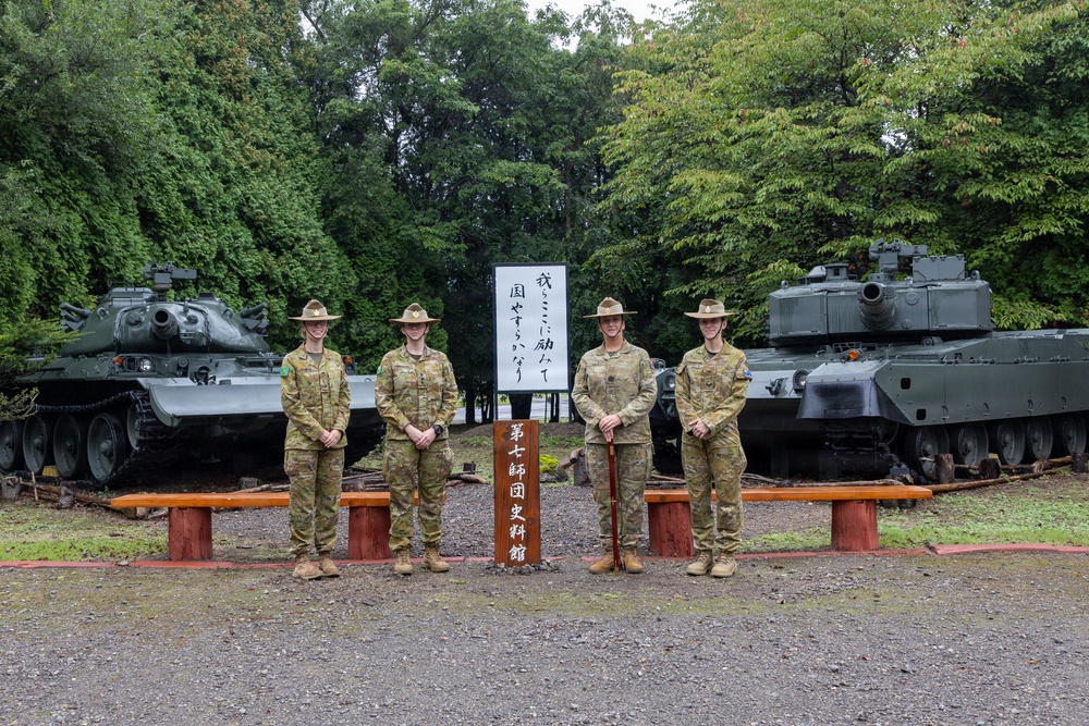 Four military personnel in Australian Army camouflage uniforms and distinctive wide-brimmed slouch hats are posing in front of a Japanese military museum sign reading '第七師団史料館' (7th Division Historical Museum). They are flanked by two static display tanks on a gravel outdoor area. The setting appears to be a Japanese Ground Self-Defense Force base, likely in Hokkaido, Japan.