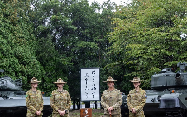 Four military personnel in Australian Army camouflage uniforms and distinctive wide-brimmed slouch hats are posing in front of a Japanese military museum sign reading '第七師団史料館' (7th Division Historical Museum). They are flanked by two static display tanks on a gravel outdoor area. The setting appears to be a Japanese Ground Self-Defense Force base, likely in Hokkaido, Japan.