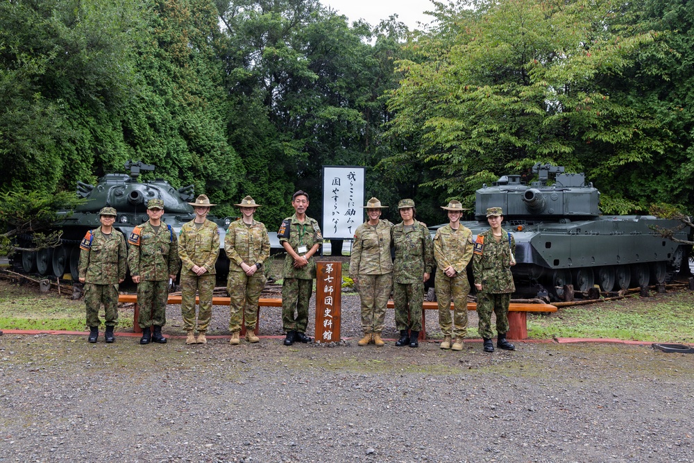 A group of nine military personnel in camouflage uniforms pose for a formal group photograph in front of a sign reading '第七師団史料館' (7th Division History Museum). Two military tanks are visible in the background, flanking the group on either side. The setting appears to be an outdoor military museum or memorial site in Japan, surrounded by dense green trees.