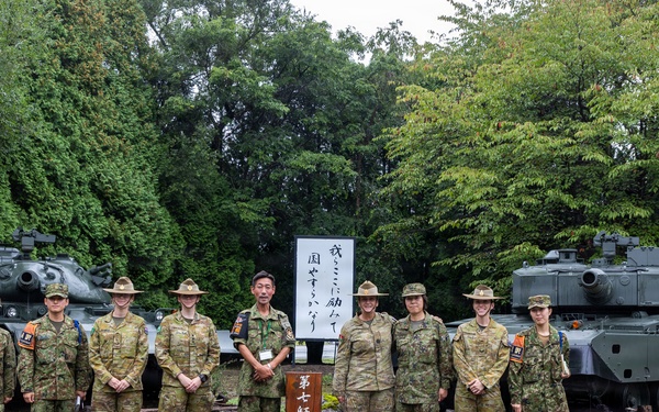 A group of nine military personnel in camouflage uniforms pose for a formal group photograph in front of a sign reading '第七師団史料館' (7th Division History Museum). Two military tanks are visible in the background, flanking the group on either side. The setting appears to be an outdoor military museum or memorial site in Japan, surrounded by dense green trees.