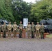 A group of nine military personnel in camouflage uniforms pose for a formal group photograph in front of a sign reading '第七師団史料館' (7th Division History Museum). Two military tanks are visible in the background, flanking the group on either side. The setting appears to be an outdoor military museum or memorial site in Japan, surrounded by dense green trees.