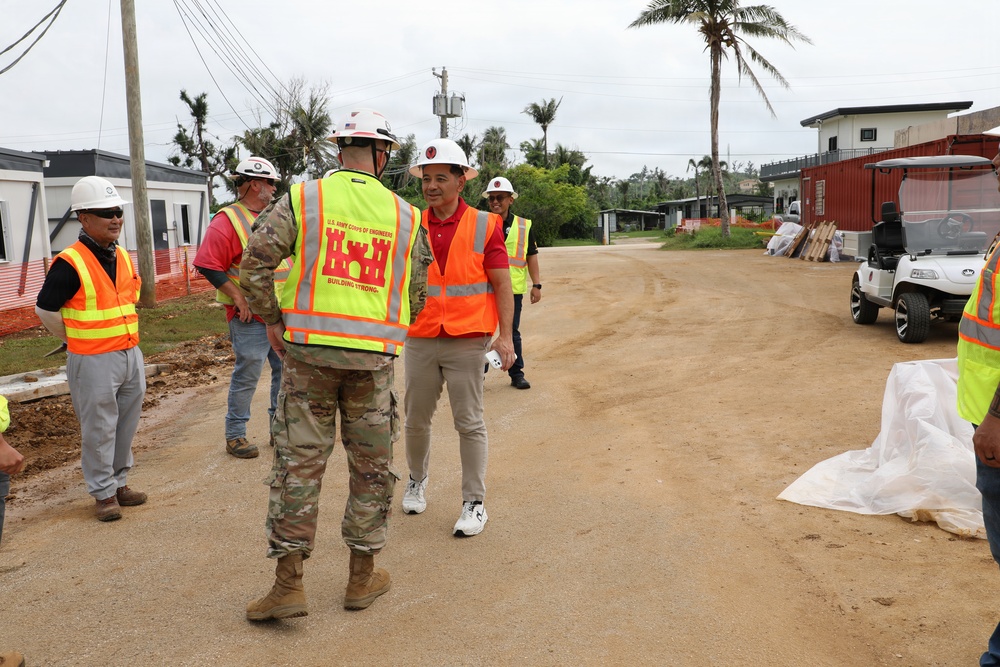 A construction site visit or inspection is underway, featuring personnel from the U.S. Army Corps of Engineers interacting with civilian officials or contractors. Multiple individuals are wearing safety gear including hard hats and high-visibility vests. The setting appears to be a tropical location, likely a U.S. territory or Pacific island, based on the palm trees and vegetation.