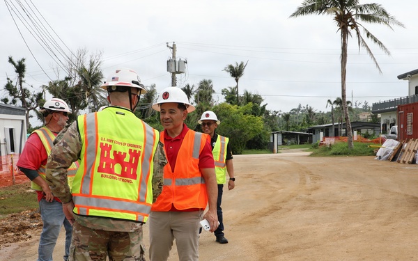 A construction site visit or inspection is underway, featuring personnel from the U.S. Army Corps of Engineers interacting with civilian officials or contractors. Multiple individuals are wearing safety gear including hard hats and high-visibility vests. The setting appears to be a tropical location, likely a U.S. territory or Pacific island, based on the palm trees and vegetation.
