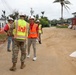 A construction site visit or inspection is underway, featuring personnel from the U.S. Army Corps of Engineers interacting with civilian officials or contractors. Multiple individuals are wearing safety gear including hard hats and high-visibility vests. The setting appears to be a tropical location, likely a U.S. territory or Pacific island, based on the palm trees and vegetation.