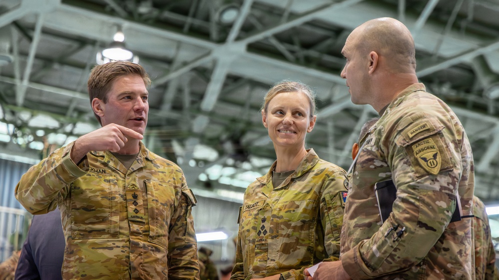 Three military personnel in camouflage uniforms are engaged in conversation inside what appears to be a military hangar or large indoor facility. Two individuals on the left wear Australian Army uniforms with 'AUSTRALIA' name tapes and rank insignia, while the individual on the right wears a US Army uniform with visible 'Ranger,' 'Airborne,' and '11th Airborne Division' patches. The interaction appears to be a professional military exchange or briefing.