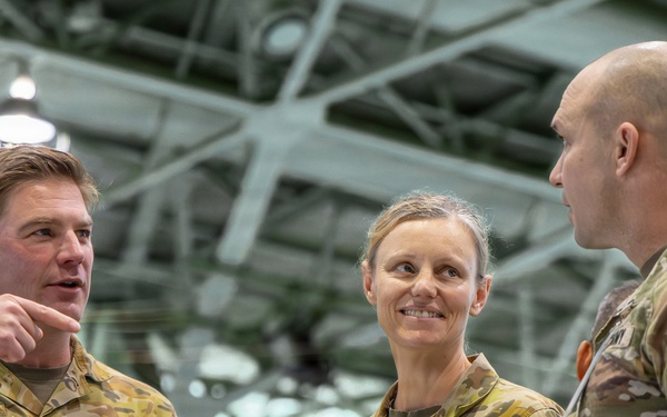 Three military personnel in camouflage uniforms are engaged in conversation inside what appears to be a military hangar or large indoor facility. Two individuals on the left wear Australian Army uniforms with 'AUSTRALIA' name tapes and rank insignia, while the individual on the right wears a US Army uniform with visible 'Ranger,' 'Airborne,' and '11th Airborne Division' patches. The interaction appears to be a professional military exchange or briefing.