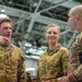 Three military personnel in camouflage uniforms are engaged in conversation inside what appears to be a military hangar or large indoor facility. Two individuals on the left wear Australian Army uniforms with 'AUSTRALIA' name tapes and rank insignia, while the individual on the right wears a US Army uniform with visible 'Ranger,' 'Airborne,' and '11th Airborne Division' patches. The interaction appears to be a professional military exchange or briefing.
