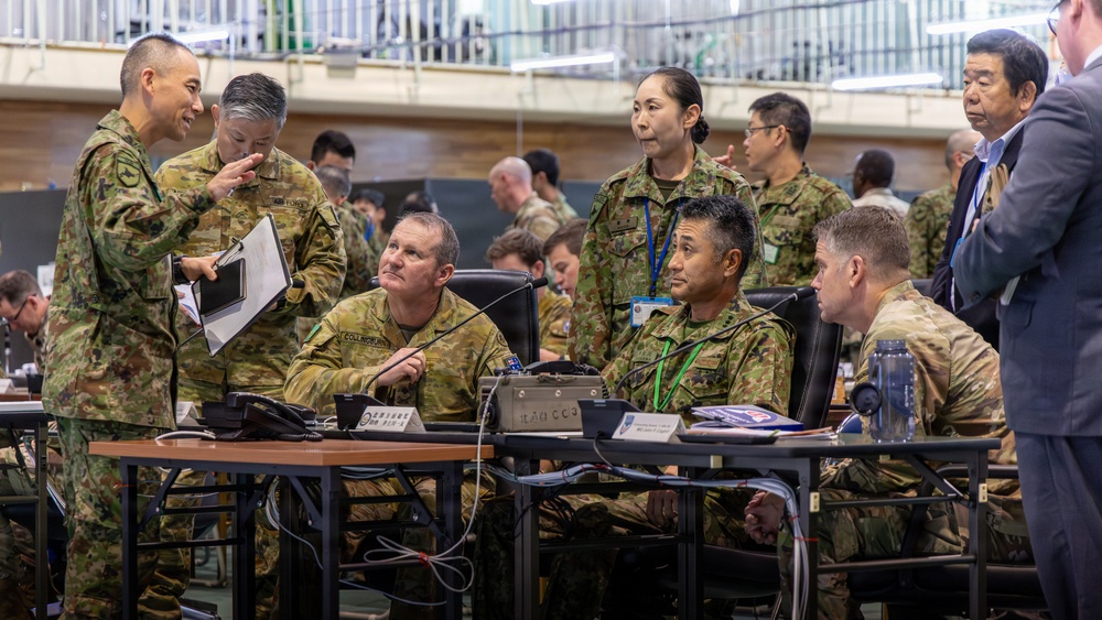 A multinational military briefing or command post exercise is taking place in what appears to be a large indoor facility. Personnel in multiple camouflage uniform patterns, including Australian Defence Force (AUSCAM) and Japanese Self-Defense Force (JGSDF) uniforms, are gathered around tables with communications equipment. At least one civilian in a suit is also present, suggesting a joint or combined military-civilian coordination event.