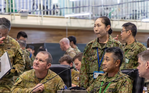 A multinational military briefing or command post exercise is taking place in what appears to be a large indoor facility. Personnel in multiple camouflage uniform patterns, including Australian Defence Force (AUSCAM) and Japanese Self-Defense Force (JGSDF) uniforms, are gathered around tables with communications equipment. At least one civilian in a suit is also present, suggesting a joint or combined military-civilian coordination event.