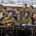 A multinational military briefing or command post exercise is taking place in what appears to be a large indoor facility. Personnel in multiple camouflage uniform patterns, including Australian Defence Force (AUSCAM) and Japanese Self-Defense Force (JGSDF) uniforms, are gathered around tables with communications equipment. At least one civilian in a suit is also present, suggesting a joint or combined military-civilian coordination event.