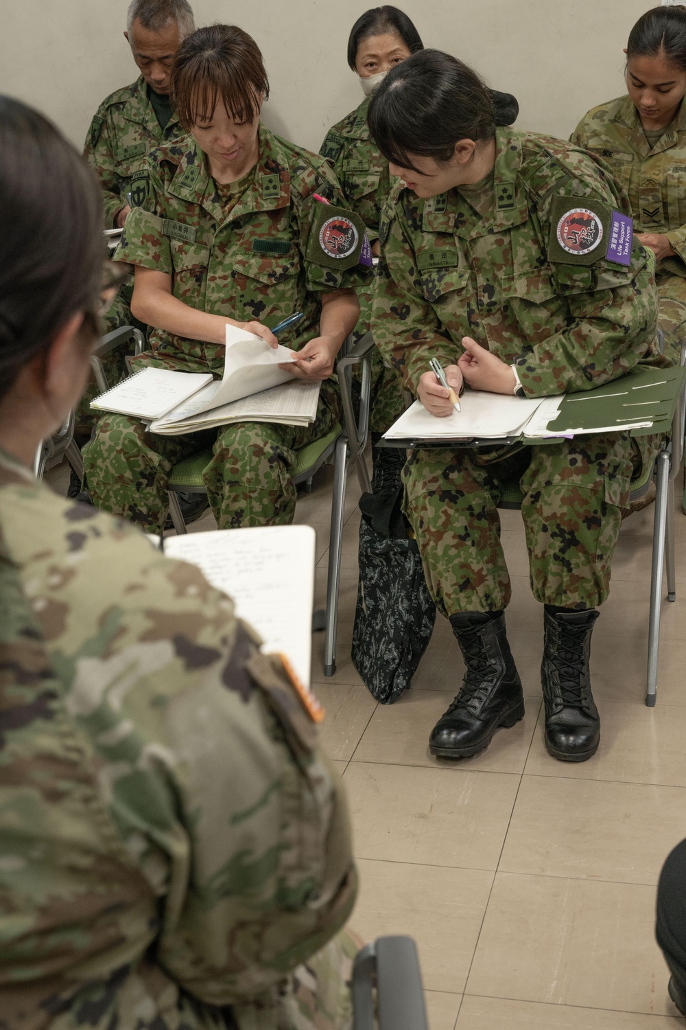 Military personnel from what appears to be the Japan Ground Self-Defense Force (JGSDF) and U.S. Army are seated in a classroom or briefing setting, taking notes on paper. The soldiers are wearing their respective camouflage uniforms — Japanese woodland digital pattern and U.S. Army OCP (Operational Camouflage Pattern). This appears to be a joint military training or coordination exercise.