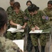 Military personnel from what appears to be the Japan Ground Self-Defense Force (JGSDF) and U.S. Army are seated in a classroom or briefing setting, taking notes on paper. The soldiers are wearing their respective camouflage uniforms — Japanese woodland digital pattern and U.S. Army OCP (Operational Camouflage Pattern). This appears to be a joint military training or coordination exercise.