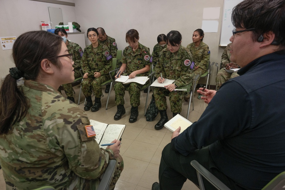 A group of military personnel from multiple nations are seated in a semicircle during what appears to be a briefing or training session in a classroom-like setting. A US Army soldier (identified by the American flag patch) and a civilian interpreter or liaison are facilitating the discussion. Several participants are taking notes in notebooks, suggesting an instructional or information-sharing exercise.