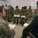A group of military personnel from multiple nations are seated in a semicircle during what appears to be a briefing or training session in a classroom-like setting. A US Army soldier (identified by the American flag patch) and a civilian interpreter or liaison are facilitating the discussion. Several participants are taking notes in notebooks, suggesting an instructional or information-sharing exercise.