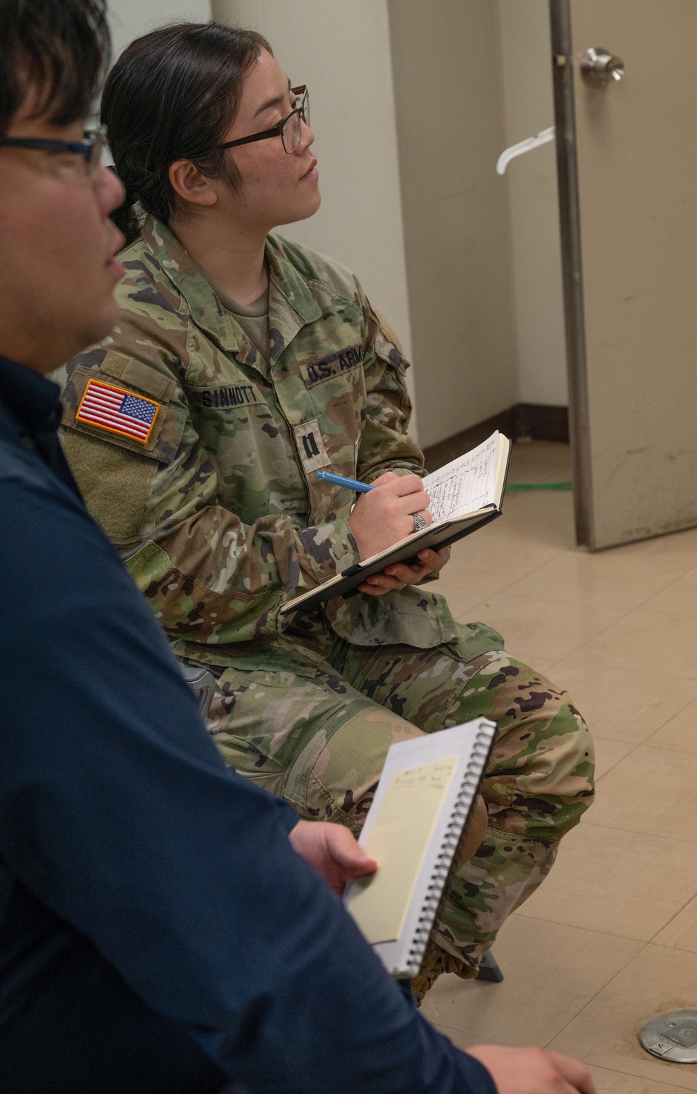 A U.S. Army soldier, identified by name tape as 'SINNOTT' and wearing the OCP (Operational Camouflage Pattern) uniform with a U.S. flag patch, is seated and taking notes in a notebook while appearing to listen attentively. A second individual in civilian clothing is partially visible in the foreground, also holding a spiral notebook. The setting appears to be an indoor facility, possibly during a briefing or training session.