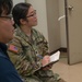 A U.S. Army soldier, identified by name tape as 'SINNOTT' and wearing the OCP (Operational Camouflage Pattern) uniform with a U.S. flag patch, is seated and taking notes in a notebook while appearing to listen attentively. A second individual in civilian clothing is partially visible in the foreground, also holding a spiral notebook. The setting appears to be an indoor facility, possibly during a briefing or training session.
