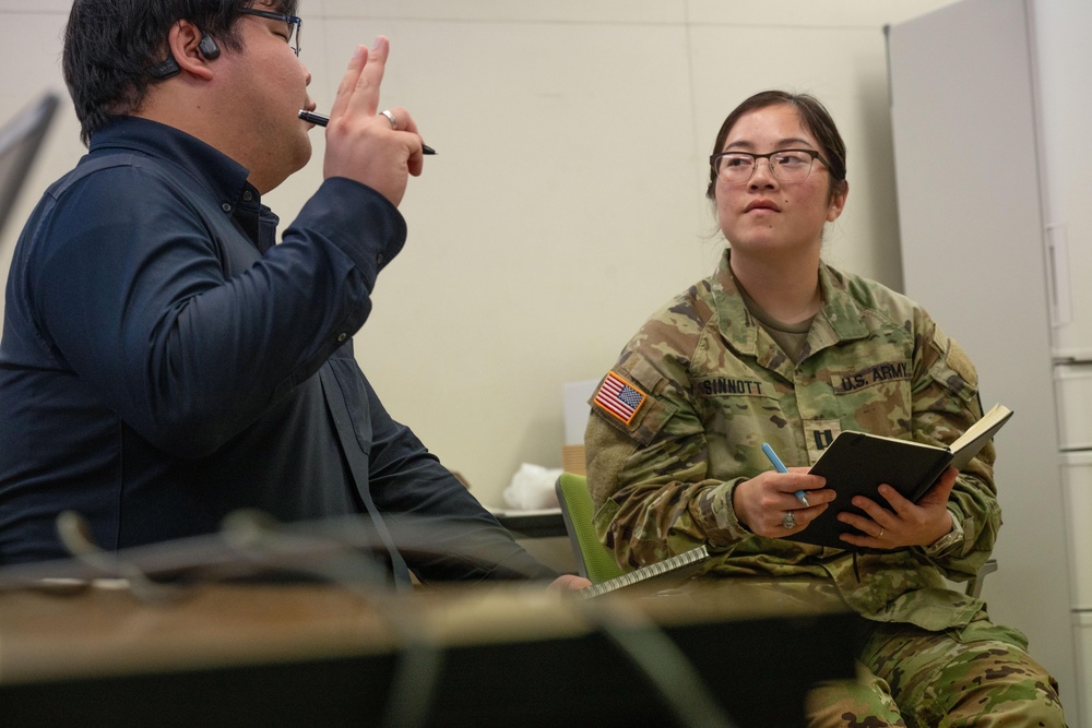 A U.S. Army soldier, identified by name tape as 'SINNOTT' and wearing an OCP (Operational Camouflage Pattern) uniform with a captain's rank insignia, is engaged in a conversation or interview with a civilian male. The soldier is holding a black notebook and a pen, appearing to take notes or conduct an interview. The setting appears to be an indoor office or meeting room environment.