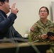 A U.S. Army soldier, identified by name tape as 'SINNOTT' and wearing an OCP (Operational Camouflage Pattern) uniform with a captain's rank insignia, is engaged in a conversation or interview with a civilian male. The soldier is holding a black notebook and a pen, appearing to take notes or conduct an interview. The setting appears to be an indoor office or meeting room environment.