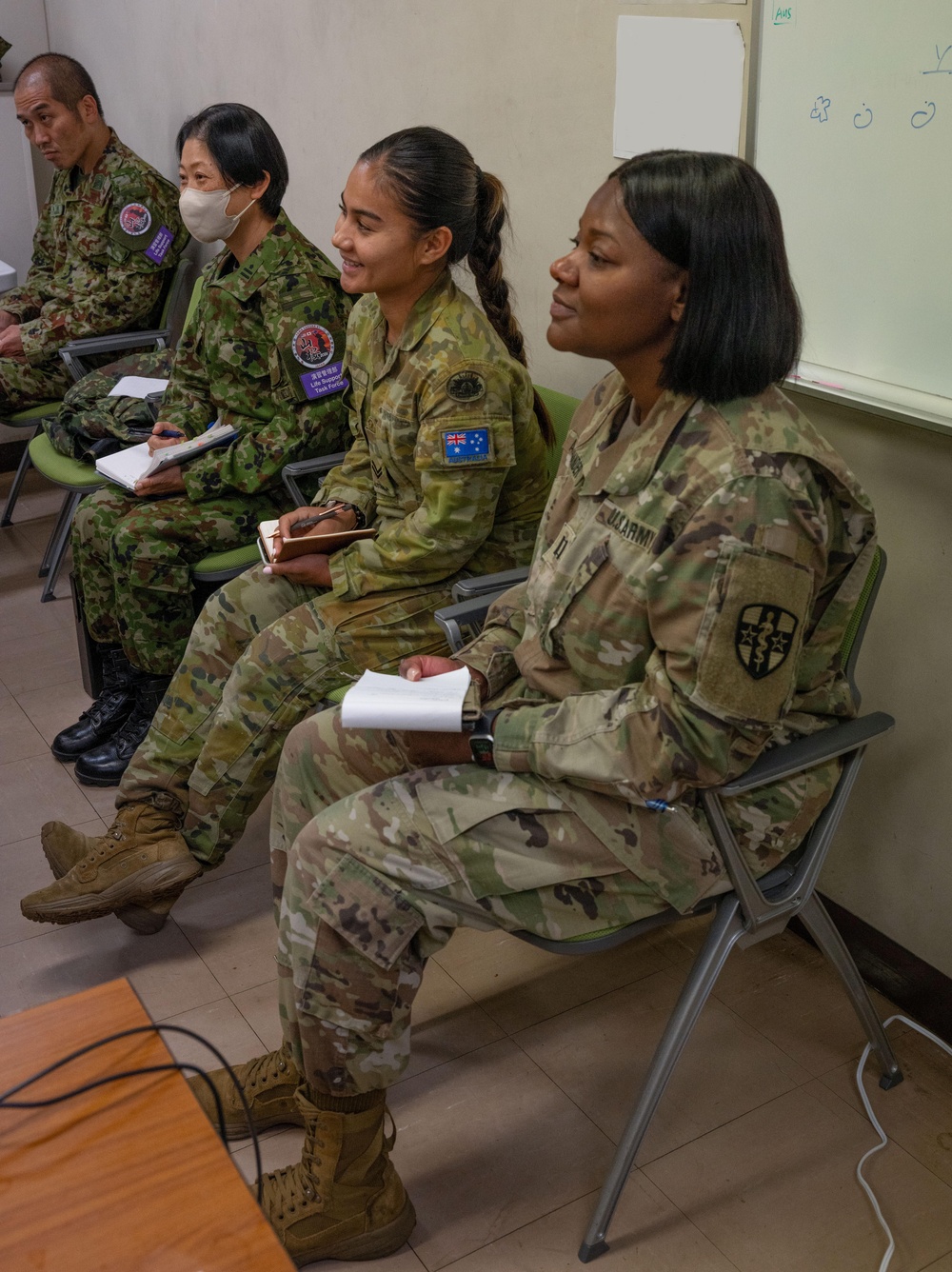 Military personnel from multiple nations are seated in a classroom or briefing room setting, appearing to attend a joint training or coordination session. At least three distinct national uniforms are visible, including Japanese Self-Defense Force, Australian Defence Force, and United States Army. Several individuals are holding notebooks, suggesting active note-taking during a presentation or lecture.