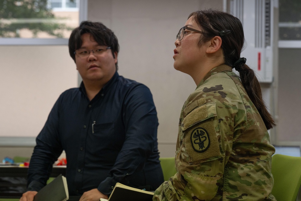 Two individuals are seated in what appears to be a meeting or briefing room, both looking in the same direction. One person is wearing a U.S. Army camouflage uniform (OCP pattern) with a Medical Corps patch on the sleeve, while the other is dressed in civilian clothing. Both wear glasses and appear attentive.